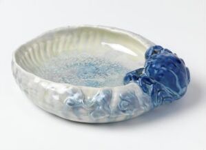  A decorative ceramic dish featuring a gradient from white to pale turquoise in the center and a bold cobalt blue sculptural adornment on the rim. The dish has a shallow, wavy edge, suggesting an organic, sea-inspired design. Artist name and title are unknown.