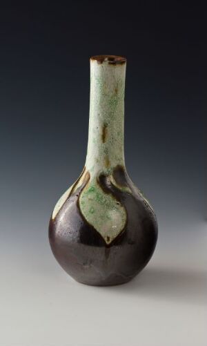  A handcrafted vase with a slender neck and rounded base exhibiting a color gradient from deep brown at the bottom to faded green at the top, standing against a neutral gray background. Artist name and title unknown.