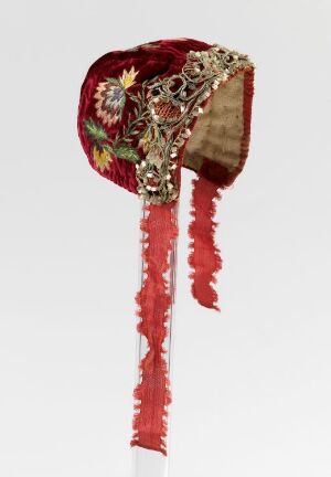  An elaborate traditional headpiece with rich red and burgundy velvet, adorned with golden floral embroidery, and frayed red ribbons flowing down.