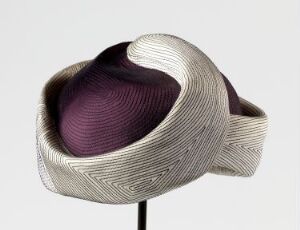  A sculptural art piece resembling a looping, twisted form similar to a stylized seashell or fantastical hat, with an intricate silvery-gray metallic exterior and a deep plum-purple interior, displayed on a stand against a neutral background.