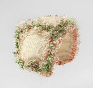  A decorative fabric cube with creamy beige base color adorned with green and pink floral lace, and edged with soft peach ruffling against a neutral background.