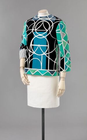  A mannequin dressed in a long-sleeve garment with a bold web-like pattern in white, black, and blue shades on a hip-length top, paired with a plain white skirt, displayed against a grey background.