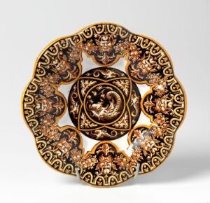  An ornately designed decorative plate featuring a deep brown and gold color scheme with baroque-inspired patterns, including a central rosette motif, acanthus leaf scrollwork, and four grotesque faces around the scalloped edges.