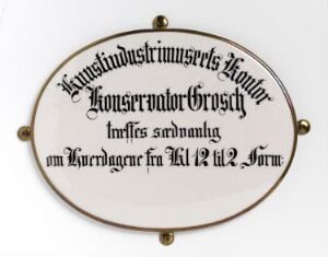  Oval-shaped porcelain plaque titled "Konservator Grosch" with a creamy white background and black stylized script, encased in a muted gold metal frame with one attachment point on each side. Created by an unidentified artist.