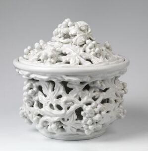  A white porcelain openwork container with a matching intricate lid, adorned with small rounded details, set against a light grey gradient background.