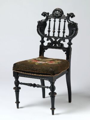  An ornately designed, black-lacquered wooden chair with intricately turned legs and backrest spindles, featuring a central relief carving and a seat with colorful floral embroidery, set against a neutral background. The work is titled "Stol" by an unknown artist.