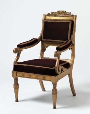  An antique armchair with golden-brown carved wooden frame and deep burgundy velvet upholstery, featuring intricately designed top crest and tapered fluted legs with metallic accents, set against a light gray background.