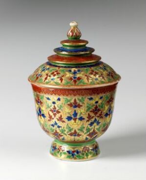  A colorful Bencharong porcelain jar with a lid, decorated with intricate floral patterns in vibrant overglaze enamel colors like blue, green, red, yellow, and white, with gold gilding accents. The jar has a