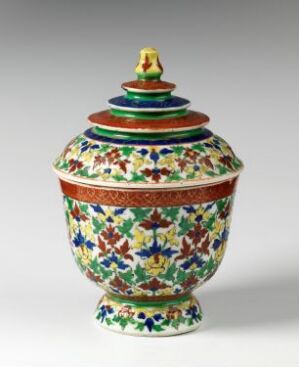  A Bencharong porcelain jar with a vibrant and intricate overglaze enamel decoration in a multitude of colors including green, red, blue, yellow and white, highlighted with gilding. The jar has a small pedestal base, a rounded body, a narrower neck, and a domed lid with a knob handle on top. Artist unknown.