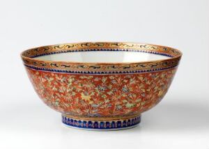  Traditional Bencharong porcelain bowl with elaborate overglaze enamel decoration in vibrant orange, adorned with blue, green, yellow, and gold motifs, showcasing a rich floral design with a gilded rim, against a soft light gray background. The bowl has a broad, curved form with a flared rim and stands on a petite rounded base.