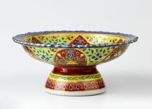  A colorful Bencharong porcelain bowl with intricate designs and gold trim, featuring a red base and a lavishly decorated exterior in shades of red, green, yellow, and blue. The bowl's interior showcases a central decorative medallion surrounded by patterned bands of various colors. Artist name: Ukjent. Title: Bencharong.