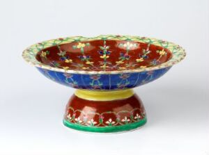  A Bencharong footed bowl made from porcelain, featuring overglaze enamel decorations in vibrant blues, reds, greens, and yellows on a white background. The artist is unknown.