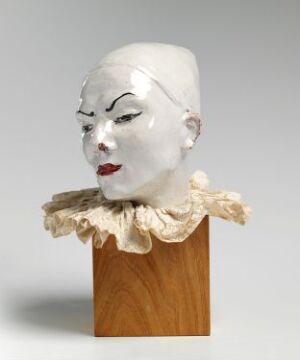  A glossy, creamy-white sculpted bust of a stylized figure with dramatic makeup and an ornate ruffled collar, set on a wooden pedestal against a light grey background. (Artist name and title unknown)