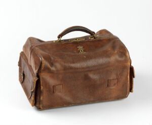  A vintage, well-used caramel brown leather doctor's bag with brass fittings and side pockets, standing against a light-colored backdrop.