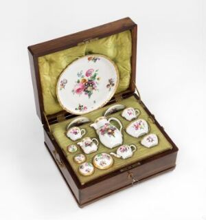  A small, open wooden box with plush yellow-green lining, containing a miniature tea set with pieces displaying floral motifs in pink, red, yellow, and blue on a white background.