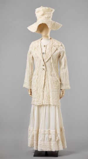  A woman's historical costume designed by Pia Myrvold, featuring a cream-colored, patterned, floor-length skirt, a fitted jacket with plastic buttons, and an elaborate wide-brimmed hat, displayed on a mannequin against a light grey background.