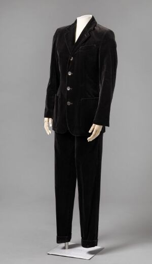  A sophisticated black velvet women's suit designed by Jean Paul Gaultier, displayed on a mannequin. It features a fitted jacket with a notched lapel and plastic buttons, and straight-legged pants, all set against a light grey background.