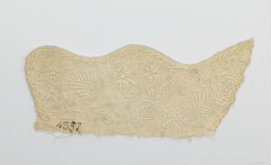  An irregularly shaped fragment of aged, off-white or cream fabric with raised floral and circular patterns, set against a solid white background, featuring a darker mark or writing in the bottom left corner. Artist name and title are unknown.