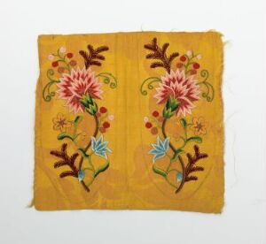  A mustard yellow square piece of fabric with symmetrical hand-embroidered floral designs in pink, red, blue, and shades of green, with frayed edges suggesting a rustic, hand-crafted textile.