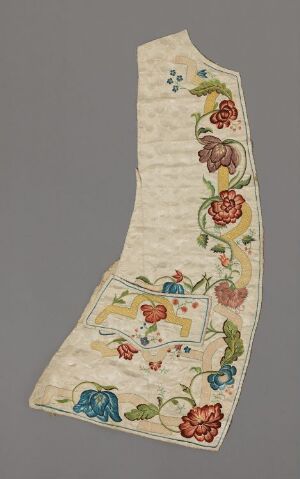  An asymmetrical, long and narrow embroidered fabric with an aged cream background, featuring vibrant, multicolored flowers connected by green vines. A detailed small scene with a house amidst the foliage is also embroidered on the fabric, with a subtle pattern of vertical rows of tiny symbols in the background.