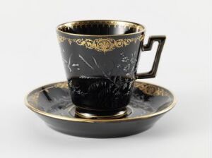  An elegant glossy black porcelain teacup with golden rim and decorative elements, paired with a matching saucer, set against a light neutral background.