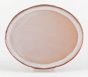  A glazed ceramic oval dish by Nina Malterud titled "Fat," featuring a muted pink-peach tone with a reddish-brown rim, set against a plain white background, illustrating the piece's gentle curves and glossy texture.