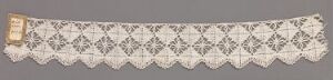  A long, intricately designed piece of white or light ivory lace fabric laid out on a grey background, featuring a net-like pattern with floral or leaf-like motifs inside diamond shapes, and a small beige label attached at one end.