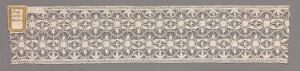  A long, narrow decorative piece with a repeating diamond and floral pattern in shades of gray on a light beige background, with a small rectangular golden emblem on the left side.