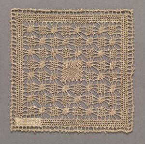  A square piece of intricately designed creamy beige lace fabric with symmetrical floral patterns, displayed against a slightly darker background.