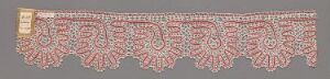  A decorative piece of lace or embroidered fabric, featuring intricate symmetrical floral motifs in shades of red and cream on a pale greyish background, with a plain segment on one edge.