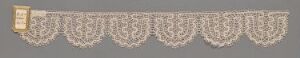  A piece of creamy white lace trim with symmetrical, intricate patterns on a grey background, attached to a golden yellow-edged tag on the left side.