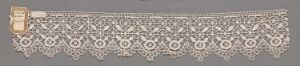  An intricate, elongated piece of white or off-white lace with a repetitive pattern of diamonds and circles that contain floral motifs, displayed against a neutral gray background with a crest or emblem attached to the left side.