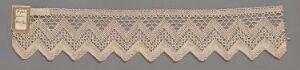  An elongated piece of intricate beige lacework with a detailed diamond pattern and a sawtooth edge, featuring a small rectangular label attached at one end, displayed against a complementary light tan background. Artist name and title are unknown.