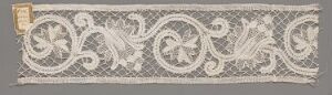  An elongated, vintage textile piece with intricate white lacework on a greyish-beige background, featuring a decorative scroll and floral pattern and a small gold-colored square label on one end.