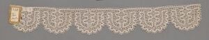  A detailed beige lace with symmetrical pattern work and scalloped edges displayed against a neutral grey background, with a square tag attached to the left side. Artist name and title are unknown.