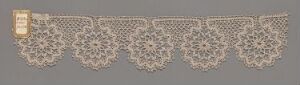  An elaborate piece of off-white lace fabric with a detailed floral pattern on a neutral grey-beige background, with a rectangular beige label attached to the left side.