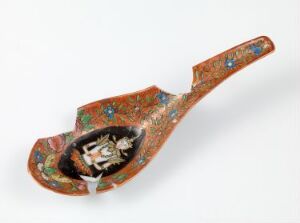  An ornately decorated Bencharong porcelain spoon with a long handle, featuring intricate enamel color designs with dominant orange and rust tones, detailed blue, green, and white floral patterns, and a central black motif with a white border in the bowl. Artist Unknown.