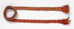  A long, red-orange woven belt or sash with a white or beige geometric pattern laid out against a light background, featuring red-orange tassels at both ends. Artist name and title are unknown.