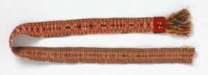  A traditional woven textile belt featuring a complex pattern in orange, red, black, white, and yellow with a solid red buckle on one end and a frayed fringe on the other, displayed against a light grey background. Artist name and title are unknown.