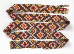  A detailed and colorful woven textile featuring a geometric pattern with red, orange, beige, and brown diamonds and crosses, highlighted with green and yellow accents against alternating dark brown and beige background segments.