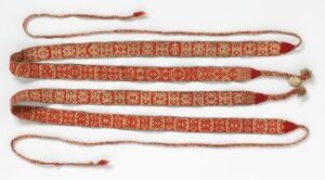  Three intricately woven red and cream textile bands parallel to each other on a neutral background, with geometric and stylized botanical designs, tassels, and a small golden ornament. Artist and title are unknown.