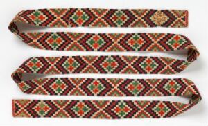  An elongated, coiled textile with a dark brown base and intricate geometric patterns in terracotta, off-white, forest green, and red, possibly a handwoven belt or strap, against a light beige background.