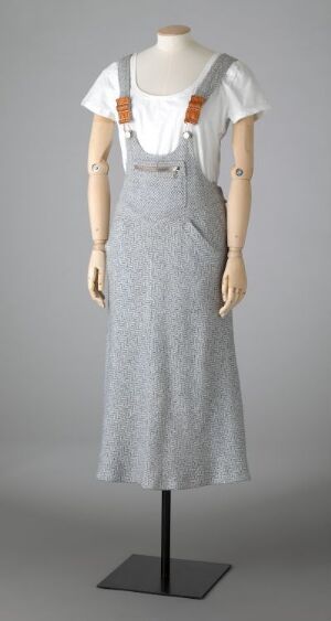  A mannequin displaying a vintage-style sleeveless gray herringbone pattern dress with brown straps and leather patches, paired with a white ruffled blouse, set against a gray background.