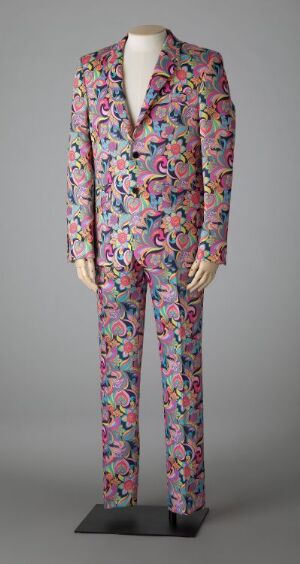  A mannequin displaying a classic men's suit with a densely packed, multicolored floral pattern on a light-toned base, featuring predominantly pink, blue, and green colors.