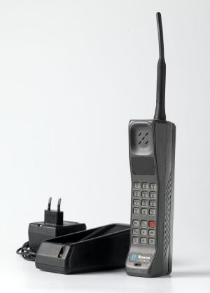  Vintage "Storno portabel" brick phone made of stainless steel and hardened plastic, shown upright with an extended antenna, next to its black charging base, against an off-white background. Artist unknown.