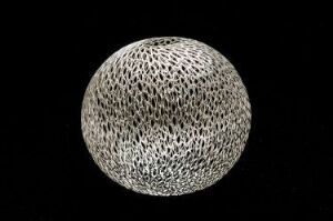  A spherical silver object with a mesh-like pattern, titled "Kule I" by artist Gunnhild Tjåland, set against a black background, showcasing the reflective and intricate design of the metalwork.