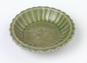  A pale green ceramic plate with a scalloped edge and a subtle embossed floral design in the center, placed on a neutral background. Artist name and title unknown.