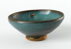  A handcrafted ceramic bowl with a deep teal glaze featuring shades of turquoise and a touch of earthy brown near the base, displayed against a plain background. The bowl has a visible small pit on its side, adding to its unique character.