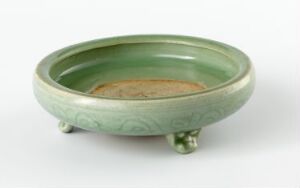  A shallow, three-footed ceramic bowl with a celadon green glaze, showing a natural beige color in the center and subtle decorative impressions beneath the glaze.