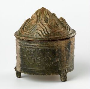  An ancient bronze container with a mountain-shaped lid and decorative relief patterns, standing on three legs against a neutral gray background.